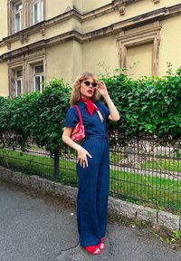 Mørkeblå linne jumpsuit med korte ærmer og bælte i taljen, suppleret med røde detaljer: et halsbånd, taske og sandaler.