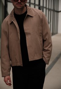 Homme portant une veste beige sur une chemise noire, avec un pantalon noir et des lunettes de soleil, debout dans un cadre urbain moderne.