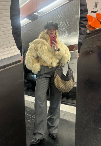 Chaqueta de piel sintética color marfil sobre una blusa marrón, pantalones grises de pierna ancha y un bolso marrón estampado. Fondo de espejo reflectante visible.