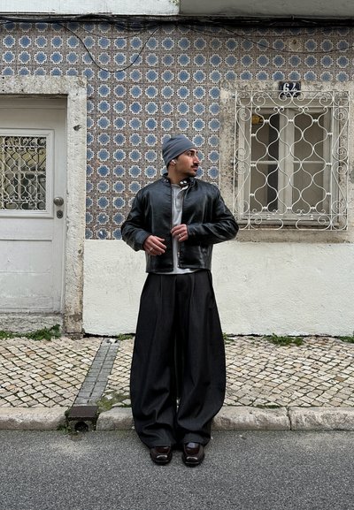 Chaqueta de cuero negra, camisa gris de gran tamaño, pantalones anchos negros y zapatos oscuros. El entorno presenta azulejos azules con patrones y una puerta blanca.