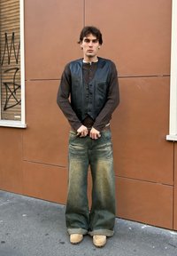 Chaleco de cuero negro sobre una camisa de manga larga marrón, combinado con unos vaqueros anchos, descoloridos y azules, y zuecos color beige sobre un fondo urbano texturizado.