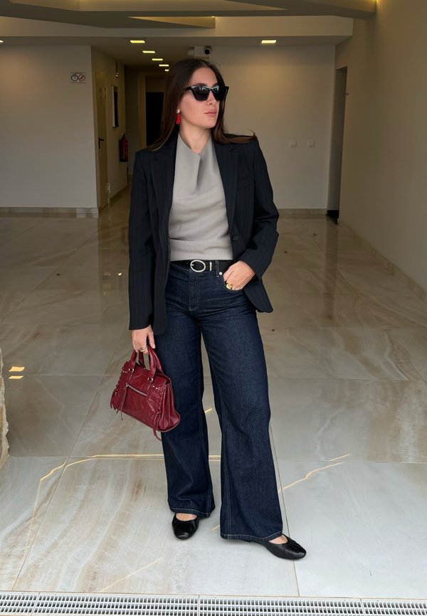 Schwarzer Nadelstreifen-Blazer, graues Top, dunkelblaue weite Jeans, schwarze Ballerinas und eine burgunderrote Handtasche. Große schwarze Sonnenbrille und rote Ohrringe.