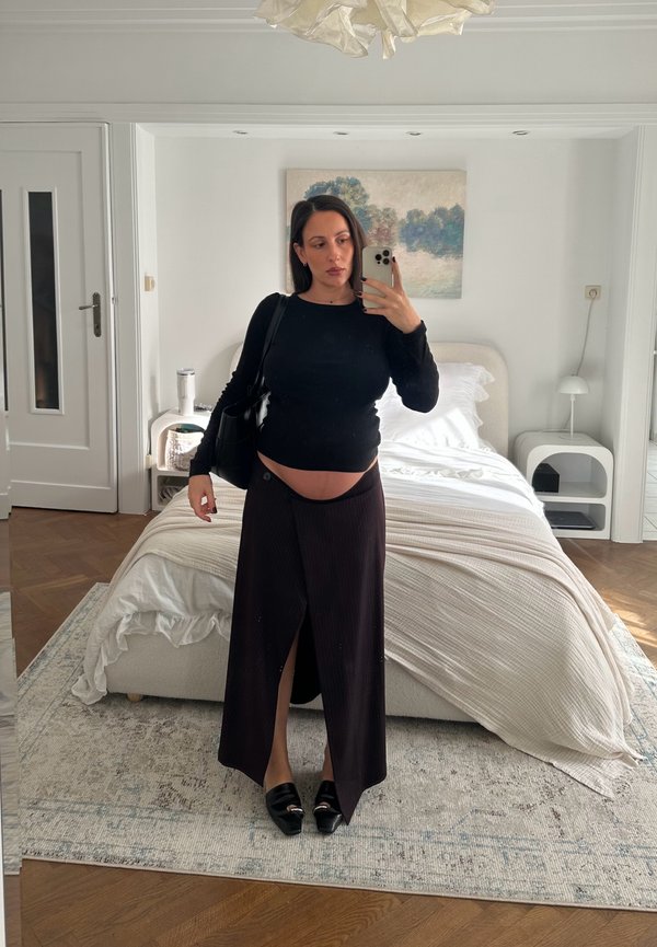 Mujer embarazada con top y falda negra se toma una selfie frente al espejo en un dormitorio moderno con cama blanca y decoración neutra.