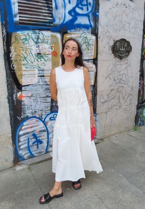 Vestido maxi de algodón blanco con diseño en capas, tirantes fruncidos y detalles con cordón. Sandalias negras de slide y un bolso de mano rojo tejido.