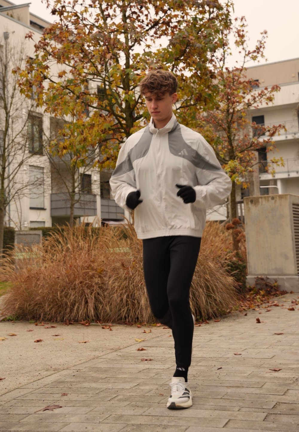 Coupe-vent blanc avec des accents gris, leggings noirs ajustés et chaussures de sport blanches. Jogging en extérieur avec un décor d'automne.