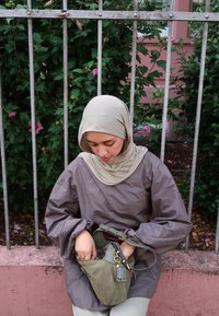 Blusa longa de mangas soltas, castanha, com mangas franzidas; hijab bege; bolsa verde-oliva com detalhes dourados. O fundo apresenta folhagem e uma parede rosa.