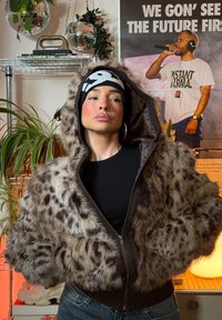Bunda z umelého kožušiny s leopardím vzorom, hnedý zips na zapínanie a rebrovaný lem. Modelka má na sebe čiernu čiapku a priliehavé čierne tričko. Vnútorné prostredie.
