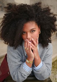 Grigia maglia lavorata a maglia con polsini a righe blu, collana di perle a strati, gioielli d'oro e capelli ricci. Mani unite davanti al viso.