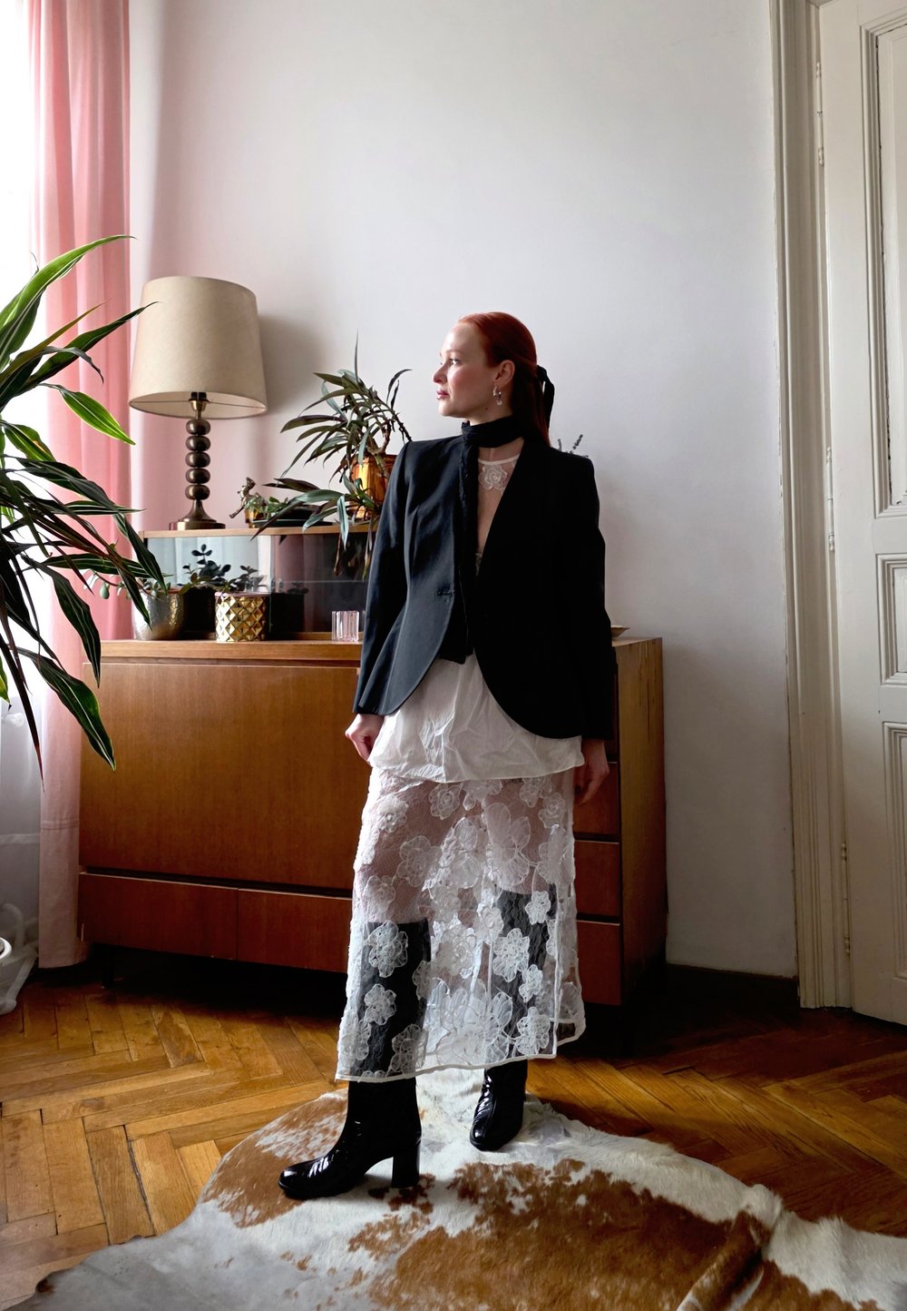 Sort blazer over en hvit blonderkjole med blomsterpreg, kombinert med blanke svarte knelang støvler. Varme tremøbler og planter i bakgrunnen.