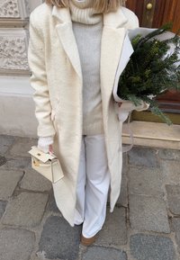 Gara, dubultsaišu mētelis teksturētā krēmkrāsā, kombinēts ar gaiši pelēku adītu augstu apkakli. Baltas bikses un neliela krēmkrāsas soma. Rokās tur zaļumu pušķi.