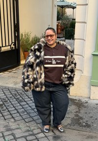 Manteau en fausse fourrure avec un motif léopard, polo marron avec des rayures claires, jean en denim foncé et chaussures grises. Debout sur un pavé en pavés.