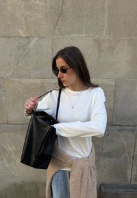 Czarna pleciona torba na ramię trzymana przez osobę w białej koszuli z długim rękawem i jasnoniebieskich dżinsach. W tle widoczna jest szara kamienna ściana o fakturze.