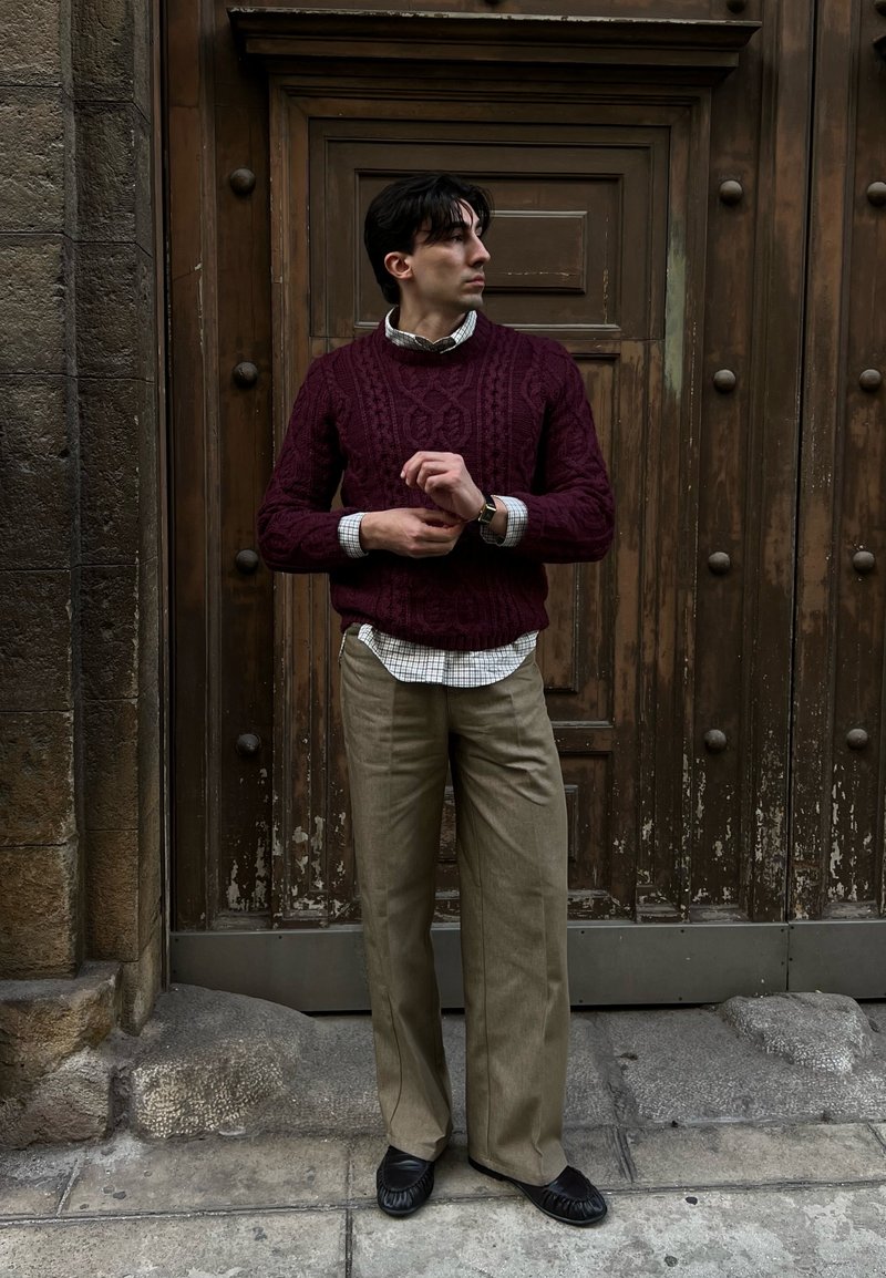 Homme debout devant une grande porte en bois, portant un pull bordeaux, une chemise à carreaux, un pantalon beige et des chaussures noires, ajustant sa montre.