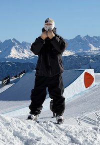 Casaco e calças de snowboard pretos, luvas bege e uma máscara facial estampada. Terreno coberto de neve e montanhas distantes ao fundo.