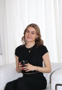 Μαύρο κύπελλο κρατημένο στα χέρια, κοντομάνικο μαύρο μπλουζάκι, χρυσές αλυσίδες, κόκκινο μανό, καθισμένος σε έναν λευκό καναπέ με λευκές κουρτίνες να κρέμονται στο φόντο.