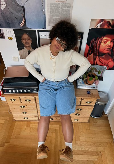 Pantalones cortos de mezclilla en azul claro, cárdigan blanco con botones, calzado marrón. Cajonera de madera, tocadiscos, planta de interior en un ambiente moderno.