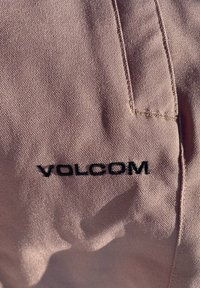 Svetlo roza tkanina z teksturirano površino, ki ima na spodnjem desnem delu izvezen črn logotip "VOLCOM". Vidne so podrobnosti šivov.