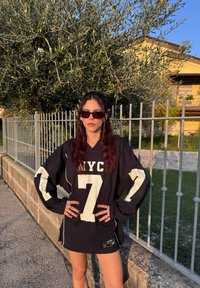 Black oversized jersey featuring white accents and the number 7, complemented by dark sunglasses. The background comprises greenery and a stone wall.