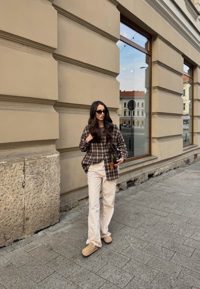 Camisa de cuadros marrón de gran tamaño, pantalones beige, calzado marrón claro y bolso negro con un llavero. El fondo presenta un edificio de color claro y pavimento.
