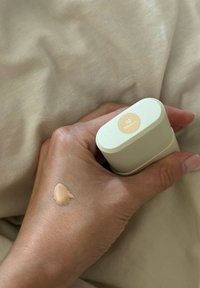 Bottiglia di fondotinta in mano che mostra una tonalità beige etichettata "02 Medio." Texture cremosa visibile sulla pelle contro uno sfondo di tessuto chiaro e testurizzato.