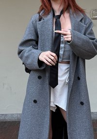 Cappotto di lana grigia con bottoni neri, indossato sopra un top corto blu chiaro e pantaloni corti bianchi texturizzati; tiene una cravatta nera.