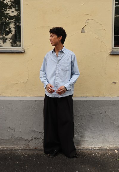 Camisa de botones a rayas azul claro, pantalones negros holgados, zapatos negros, de pie contra una pared amarilla texturizada con una ventana.