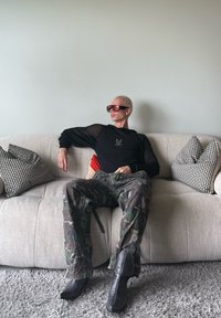 Zwarte lange mouwen mesh top, camouflage cargo broek en puntige zwarte laarzen. Zittend op een beige bank met patroonkussens. Grijs tapijt.