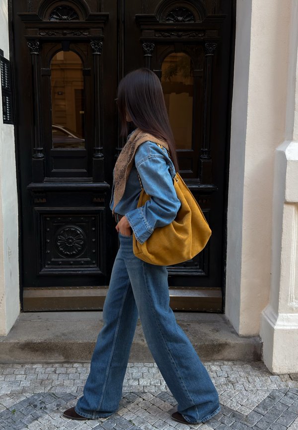 Une femme vêtue de denim et portant une écharpe passe devant une porte noire ornée, un grand sac jaune moutarde à l’épaule.