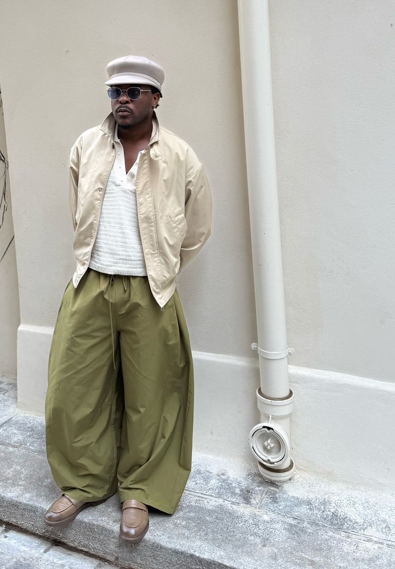 Veste bomber beige portée sur un haut en maille blanche, associée à un pantalon large vert olive et des mocassins marron. Accessoirisée d'une casquette gris clair et de lunettes de soleil.