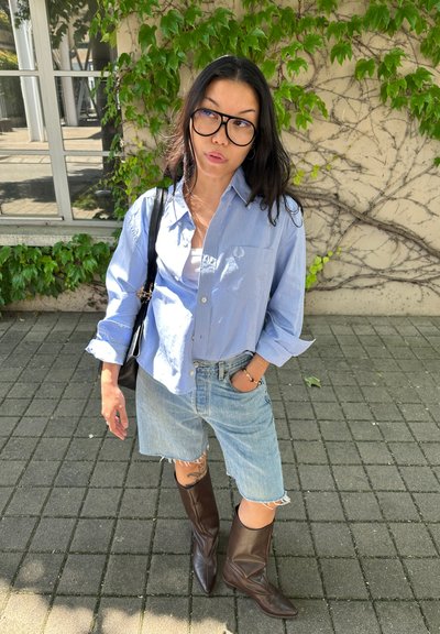 Camisa de botones azul claro con un bolsillo, combinada con pantalones cortos de mezclilla desgastados. Botas marrones de punta afilada hasta la rodilla completan el conjunto.