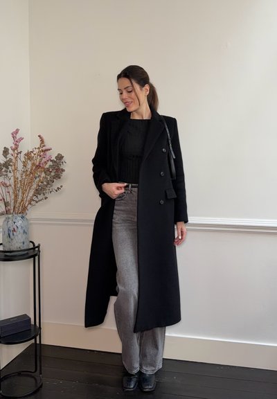 Mujer de pie en interiores, vestida con un abrigo largo negro, camiseta negra, vaqueros grises y botas negras, al lado de una mesa pequeña con flores secas.