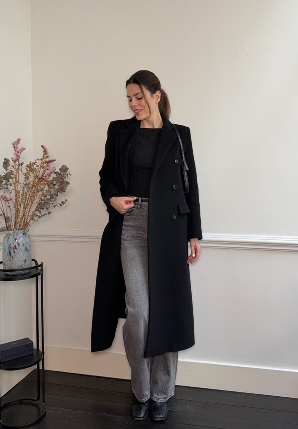 Kobieta stojąca w pomieszczeniu, ubrana w długi czarny płaszcz, czarną bluzkę, szare jeansy i czarne buty, obok małego stolika z suszonymi kwiatami.