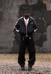 Homme aux cheveux tressés portant une veste noire, une cravate rose, un pantalon noir avec des porte-clés, debout sur du gravier devant un mur sombre et usé.