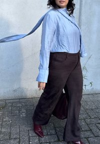 Blusa azul clara com um laço, mangas longas, combinada com calças flare castanhas escuras e sapatos de salto agudo burgundy, contra um fundo texturizado.