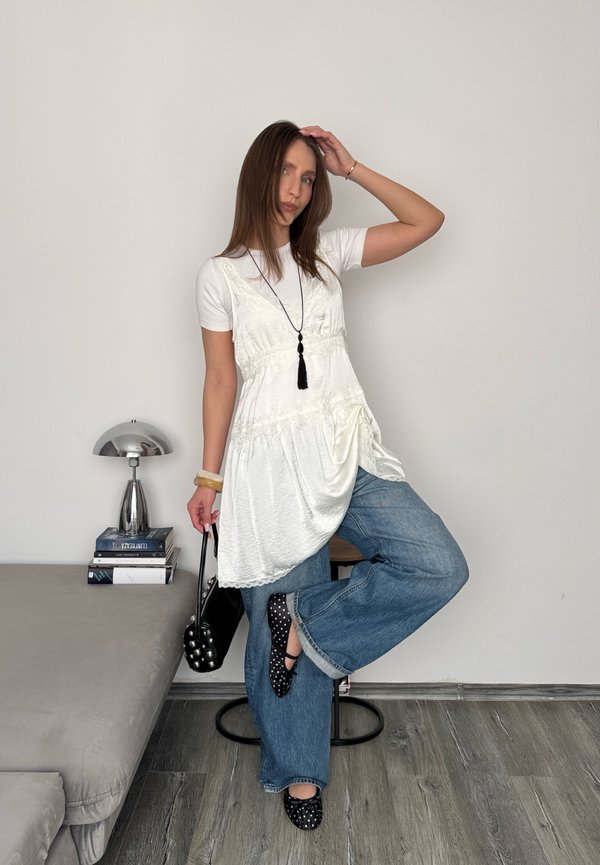Kvinne i hvit bluse, løse blå jeans og svarte sko med prikker står på ett ben og holder en svart håndveske i et minimalistisk rom.
