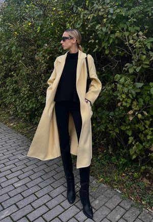 Largo abrigo de lana color crema con frente abierto, usado sobre un suéter negro y pantalones vaqueros negros. Botas de tacón negras hasta la rodilla completan el conjunto.