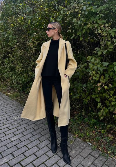 Largo abrigo de lana color crema con frente abierto, usado sobre un suéter negro y pantalones vaqueros negros. Botas de tacón negras hasta la rodilla completan el conjunto.