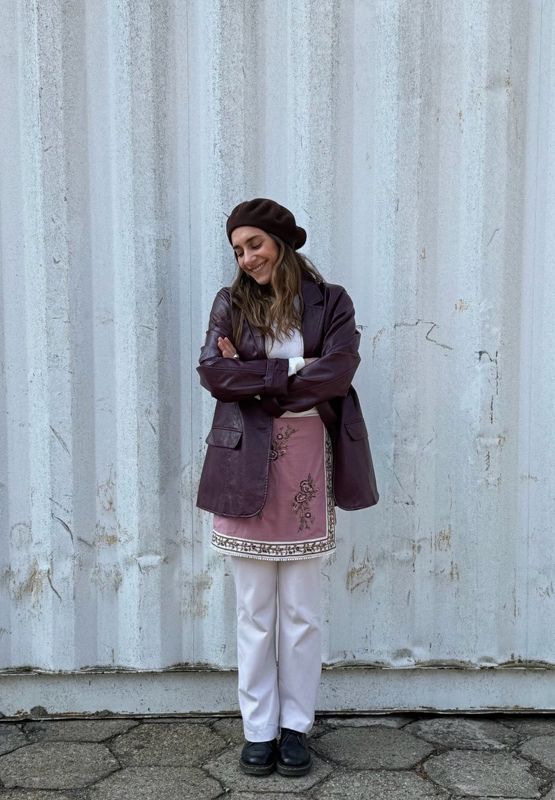 Noor naine, kellel on pikad juuksed ja kes kannab pruunikat baretti, tumelillat jopet, tikandiga roosat põllseelikut, valgeid pükse ning musti kingi, seisab käed rüpes.