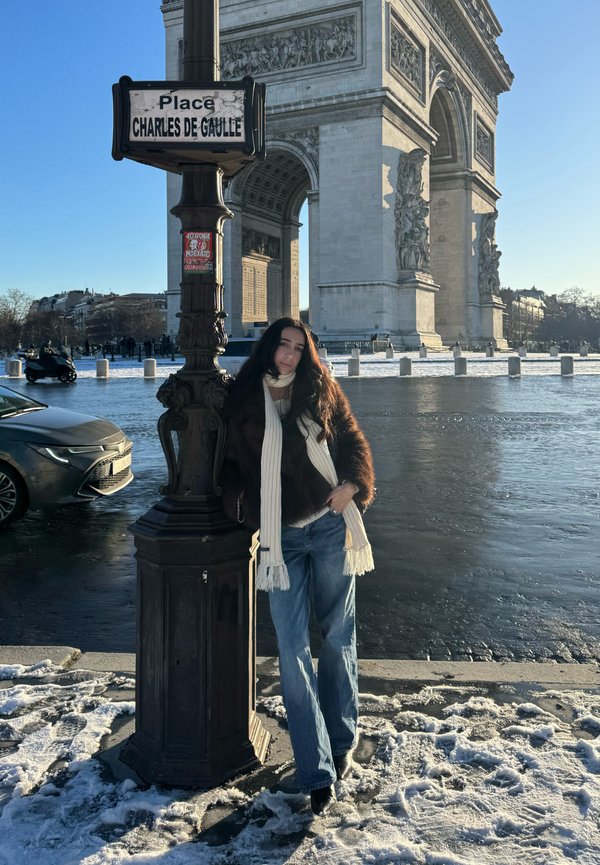 Femeie îmbrăcată în haine de iarnă sprijinindu-se de un stâlp cu indicatorul stradal "Place Charles de Gaulle" lângă Arcul de Triumf, pe o stradă din Paris acoperită de zăpadă.