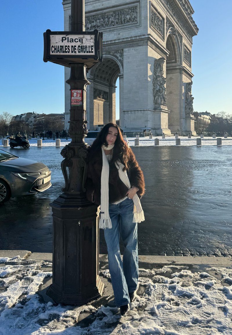 Vrouw in winterkleding die leunt tegen een straatnaambordpaal met de tekst "Place Charles de Gaulle" nabij de Arc de Triomphe op een besneeuwde straat in Parijs.