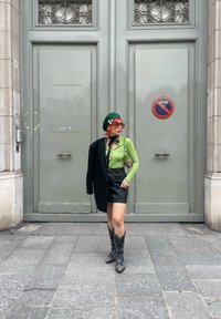Modelo vestindo uma blusa verde de mangas longas, saia preta e botas de cowboy, segurando um blazer risca de giz com uma bolsa estampada e óculos de sol vermelhos.