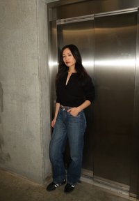 Black button-up shirt, loose blue jeans, and black sneakers with white stripes. Standing next to a metallic lift in a concrete environment.