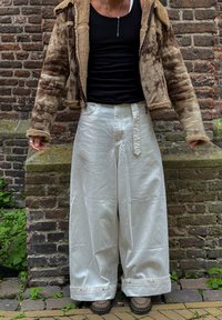 Beżowa kurtka z futerkiem z kapturem, czarny top na ramiączkach oraz blady biały blezer z szerokimi nogawkami o błyszczącej teksturze i wyeksponowanej talii.