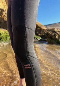 Gros plan sur une personne portant un pantalon de combinaison noire debout dans une eau côtière peu profonde et rocheuse sous un ciel bleu clair.