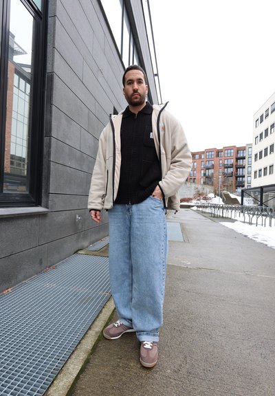 Hombre con chaqueta beige, suéter negro, vaqueros azul claro y zapatillas marrones parado en una acera de la ciudad cerca de edificios y aparcamientos para bicicletas.