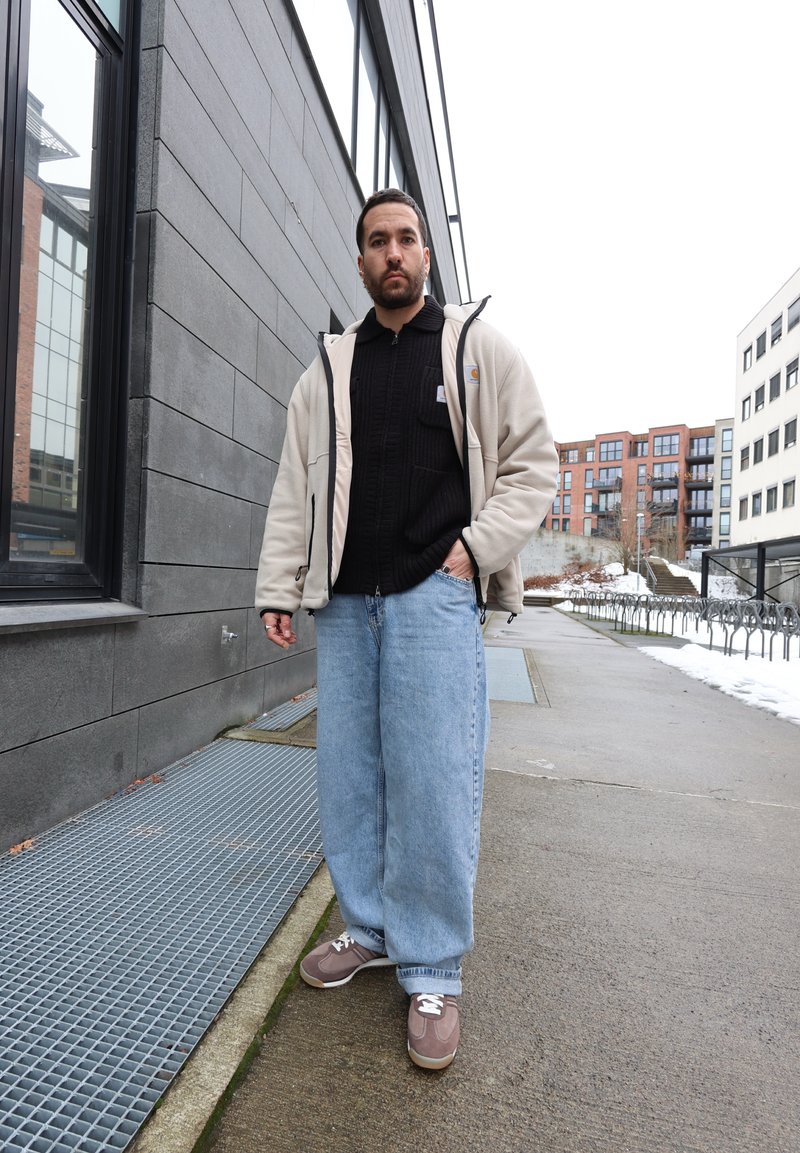 Mann iført beige jakke, svart genser, lyseblå jeans og brune joggesko står på en fortau i byen nær bygninger og sykkelstativer.