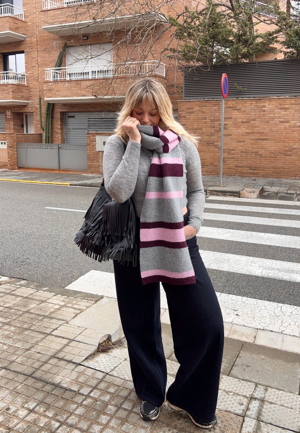 Femme blonde portant un pull gris, une écharpe rayée, un pantalon large noir et des baskets, debout près d'un passage piéton tenant un sac noir à franges.