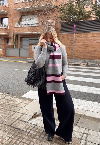 Mujer rubia que lleva un suéter gris, bufanda a rayas, pantalones anchos negros y zapatillas, de pie cerca de un paso de peatones sosteniendo una bolsa negra con flecos.