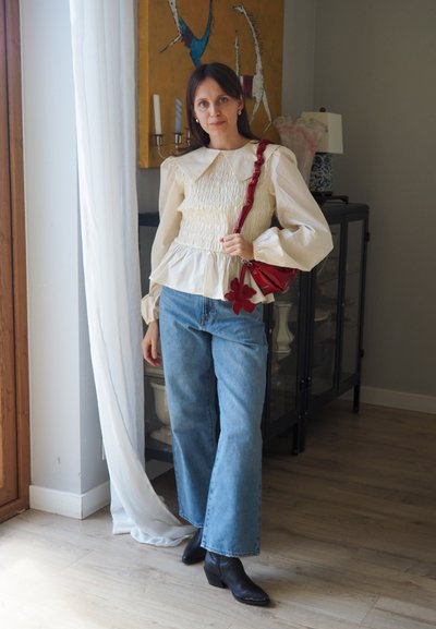 Blusa blanca de manga larga recogida con un gran cuello; jeans de pierna ancha de color azul claro; botines negros; bolso rojo con un acento de flor. Interior minimalista.