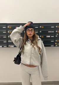 Grå hettegenser med glidelås og hette, kombinert med grå leggings. Svart strikke lue, svart håndveske og runde briller. Postkasser i bakgrunnen.